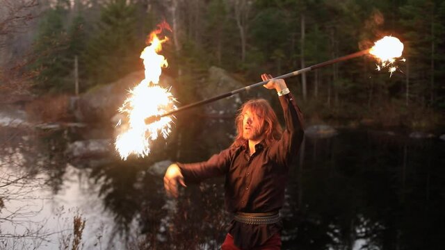 Handsome young male performer performing fire pois with fire stick standing near lake in forest and hitting flames as performance during fire jamming