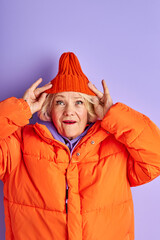 elderly lady in orange warm clothes posing at camera isolated over purple background, fancy stylish woman in shock, stand with wide opened eyes