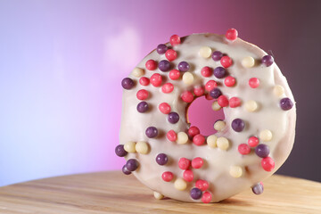 White icing donut with small colored pieces of chocolate on a wooden table against purple background