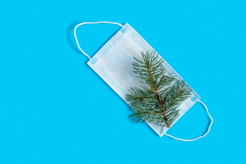 A sprig of a Christmas tree on a protective white mask on a blue background.Christmas, new year during the coronavirus pandemic, the COVID holiday. Safety during the winter Covid-19 holidays. Top view