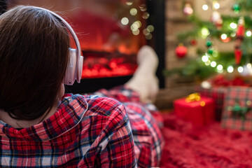 Woman in pajama in headphones lying listening music and warming at winter evening near fireplace...