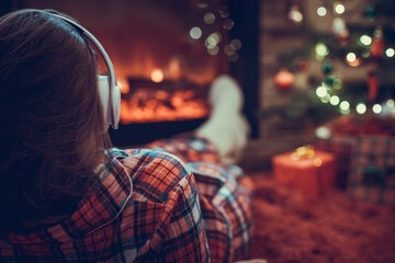 Fototapeta na wymiar Woman in pajama in headphones lying, relaxing and warming at winter evening near fireplace flame christmas tree.