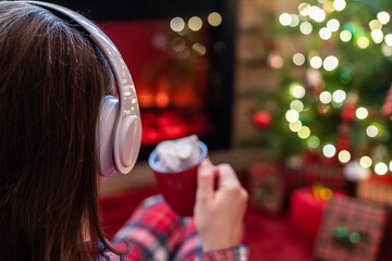 Woman in pajama in headphones with cup of hot cocoa and marshmallow sitting and warming at winter...