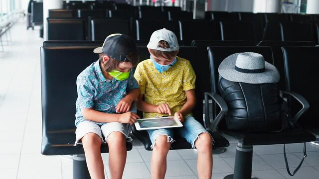 Masked children use tablet in empty airport lounge, new normality during travel. Obligation to wear face mask indoors in public places.