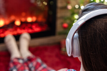 Woman in pajama in headphones sitting and warming at winter evening near fireplace flame and ...