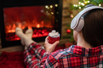Woman in pajama in headphones with cup of hot cocoa and marshmallow sitting and warming at winter...