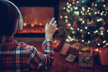 Alone sad woman in headphones sitting and warming at winter evening near fireplace flame and  christmas tree.