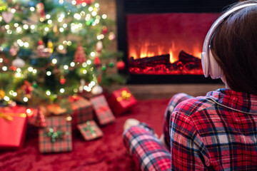 Woman in pajama in headphones sitting and warming at winter evening near fireplace flame and ...