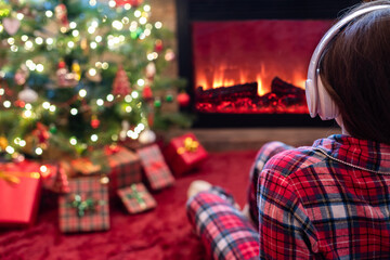 Woman in pajama in headphones sitting and warming at winter evening near fireplace flame and  christmas tree.