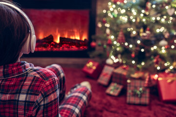 Obraz premium Alone sad woman in headphones sitting and warming at winter evening near fireplace flame and christmas tree.