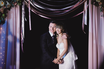 Fashionable newlyweds stand at night against the backdrop of chic decorations and flowers,electric bulbs and garlands,in the backlight. Night ceremony. Floristics. Grain,film effect