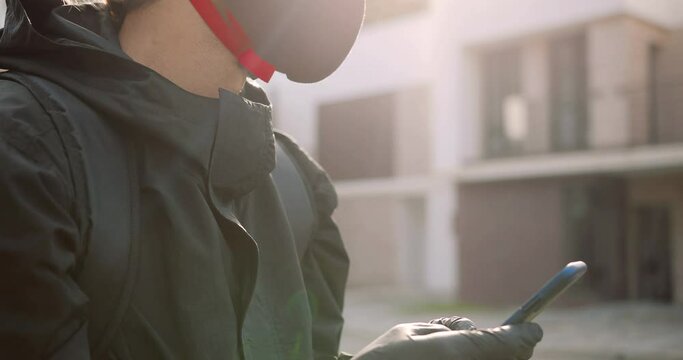 Close up of young Caucasian male courier in black jacket on street with thermal bag texting on smartphone. Handsome delivery man typing on cellphone while waiting for client. Delivery concept