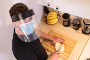 Close up shot from above of a young man in the kitchen, chopping an onion on a cutting boar wearing covid protective face mask and plastic visor