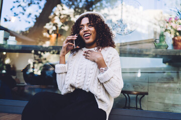 Cheerful african american woman in trendy wear sitting on bench on street making mobile phone on free time, smiline dark skinned female satisfied with international connection talking on smartphone