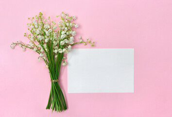 White flowers Lily of the valley ( Convallaria majalis, May bells, may lily ) on a pink paper background with white paper card note with space for text. Top view, flat lay. Spring decoration
