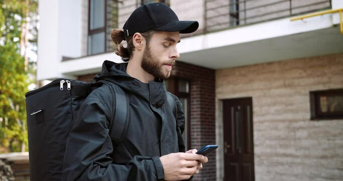 Side view of young male employee with big backpack walking on street in city and texting on smartphone looking for direction. Caucasian delivery man typing on cellphone searching for address. Close up