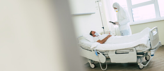 An adult male patient lies on a bed in a hospital ward while he is examined by a doctor in protective clothing and a mask.