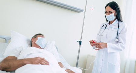A confident doctor woman in a safety medical mask is examining a mature patient in the mask while he lying in the ward.