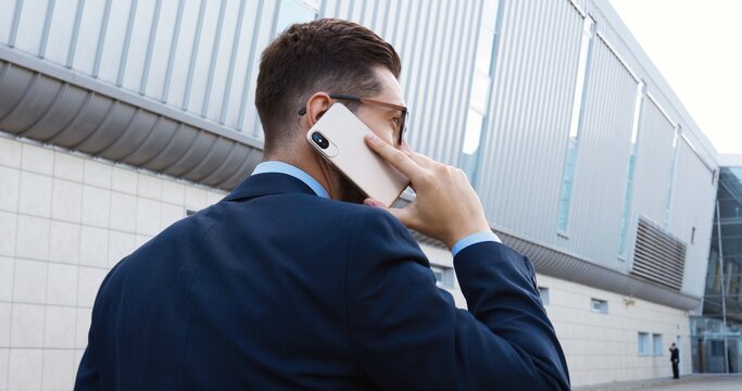 Rear of Caucasian handsome young businessman in suit, tie and glasses talking on smartphone and walking outdoors. Back view on good-looking man in business style speaking on mobile phone.