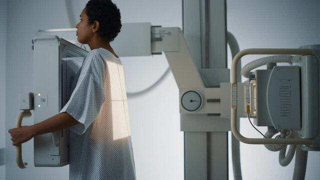 Hospital Radiology Room: Beautiful Latin Woman Standing while Professional Female Radiologist Explains Procedure, Adjusts X-Ray Machine. Scanning Chest, Heart, Lungs. Mammogram Cancer Screening