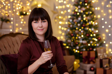 young beautiful woman drinking champagne in decorated living room with Christmas tree and festive led lights