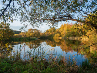 Lake surrounded by colorful trees in golden autumn evening. Blue sky and bright warm colors of leaves reflecting on water surface.