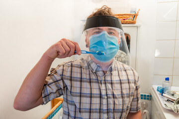 Funny and ironical shot of a young man in his bathroom trying to wash his teeth with and over a covid protective face mask and plastic visor. - obrazy, fototapety, plakaty