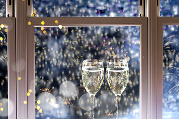 New Years background with two glasses of champagne, falling snow and firework display viewed by window. 