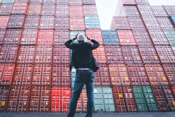 Worker or foreman standing in front of stacked cargo containers and hands hold his head wears white...