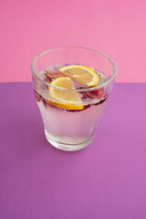 a glass of sparkling water cocktail with sliced lemon, cherry and berry. non-alcoholic drink, beverage. purple textured backdrop and pink background. carbonated water and bubble. abstract, close up.