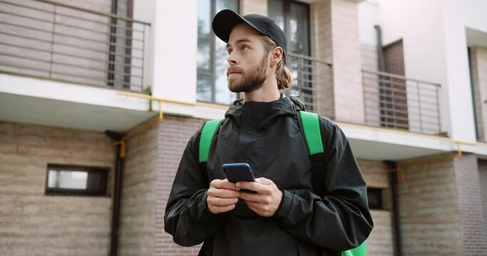 Portrait of young male employee with big backpack standing on street and texting on smartphone looking for direction to deliver orders. Caucasian delivery man typing on cellphone searching for address