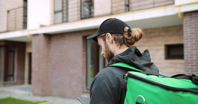 Close up of Caucasian male worker with big green thermal bag walking on street in city looking for address to deliver order. Delivery young man walk in town. Back view. Supply concept