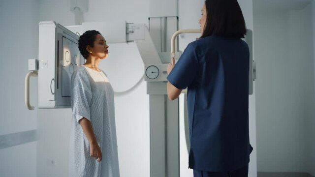 Hospital Radiology Room: Beautiful Latin Woman Standing while Professional Female Radiologist Explains Procedure, Adjusts X-Ray Machine. Scanning Chest, Heart, Lungs. Mammogram Cancer Screening