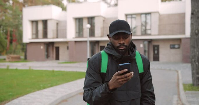 Portrait of young African American male employee walking on street and texting on smartphone looking for direction to deliver orders. Delivery man typing on mobile phone. E-commerce concept