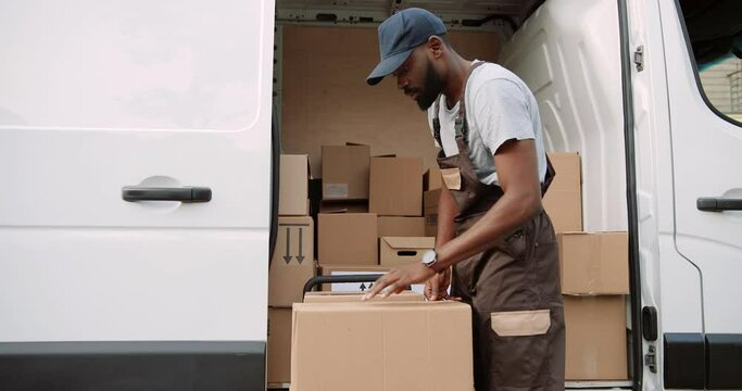 Young happy African American delivery man taking box from van standing on street outdoors and smiling at camera. Joyful male postman with ordered shopping package. Shipment concept