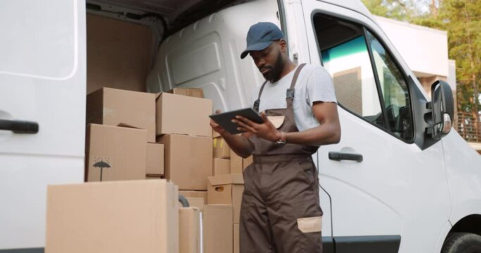 Young handsome African American delivery man typing and tapping on tablet device standing near van on street with many ordered boxes. Male postman courier with shopping package. Shipment concept