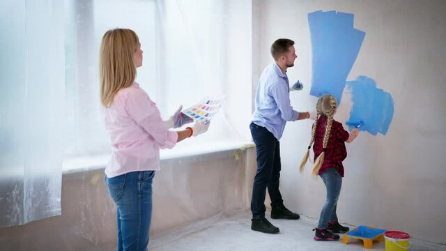 house room renovation, young family are showing how to paint walls with rollers that are covered in blue paint to their adorable small daughter