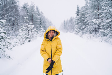 Fototapeta na wymiar Half length portrait of happy female in winter clothes putting on gloves spending time on rural white snowy road,smiling woman explorer standing in wood with tall frozen trees