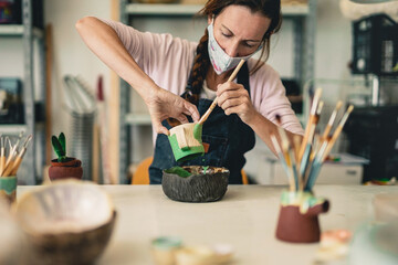 Caucasian woman works with ceramics and paints finished cup while wearing protective face mask for coronavirus - Art work studio
