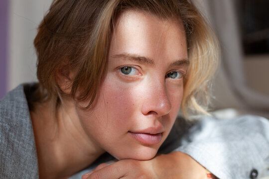 Close-up portrait of beautiful young blonde woman without make-up in gray linen shirt in interior with sunlight