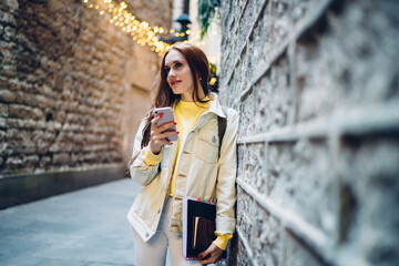 Pretty female blogger with education equipment in hand standing near wall and smiling during mobility time for online socialising, attractive red hair hipster girl holding smartphone gadget