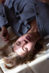 Obraz na płótnie Canvas Close-up portrait from top of beautiful young blonde woman without make-up in gray linen shirt is lying on sofa in interior with sunlight