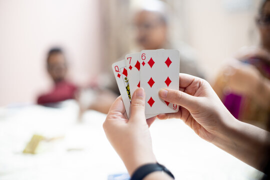 Woman Holding Cards With Out Of Focus Family Members In The Distance Showing A Family Playing Cards On Festivals Like Diwali, Dussera, New Year And More