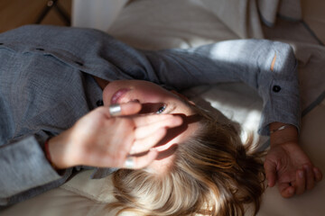 Fototapeta premium Close-up portrait from top of beautiful young blonde woman without make-up in gray linen shirt is lying on sofa in interior with sunlight