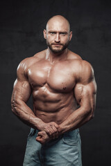 Fototapeta na wymiar Muscular and bald caucasian man with perfect abs and huge biceps poses in dark background looking at camera with serious face.