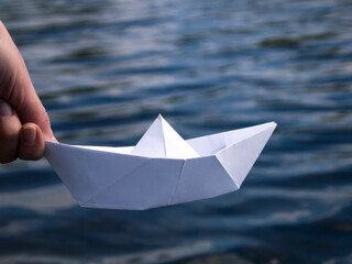 A hand is holding a white paper boat above the water. Origami ship - symbol of new beginnings. The...