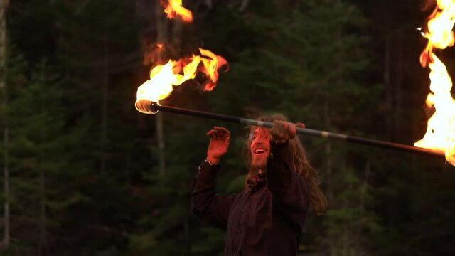 Excited young male performer performing fire pois with fire stick standing near lake in forest and hitting flames as performance during fire jamming