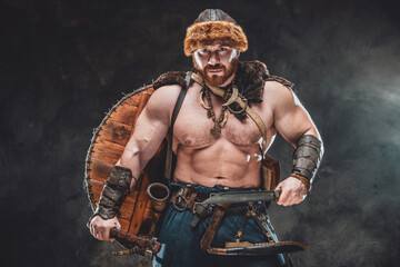 Fototapeta na wymiar Portrait of shirtless muscular viking with shield on his back and armed with hatchet and knife he poses on dark foggy background.