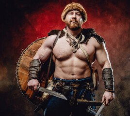 Fototapeta na wymiar Armed with two knives muscular and shirtless northern barbarian with hat and shield on his back poses in dark red background.