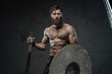 Obraz na płótnie Canvas Furious medieval nord warrior with muscular build and naked torso posing in dark background holding his helmet and axe.